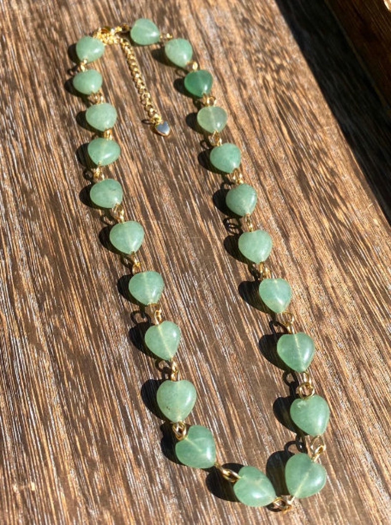 Pu&ograve; includere: Una delicata collana con pietre preziose verdi a forma di cuore infilate su una catena color oro. La collana ha un piccolo ciondolo a forma di cuore sulla chiusura, esposta su una superficie di legno.