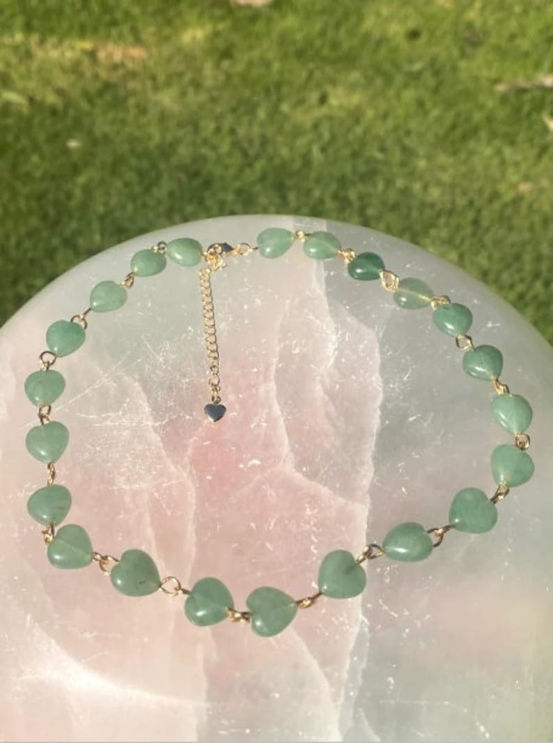 Pu&ograve; includere: Una delicata collana con pietre preziose verdi a forma di cuore infilate su una catena color oro. La collana ha una lunghezza regolabile e un piccolo ciondolo a forma di cuore sulla chiusura. La collana &egrave; esposta su una superficie rosa chiaro.