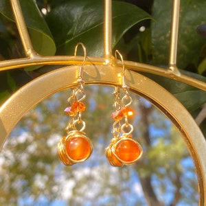 May include: A pair of handcrafted gold-colored wire-wrapped earrings. Each earring features a round, orange-colored gemstone encased in gold wire, with smaller orange crystals and gold chain accents.