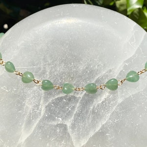 Pu&ograve; includere: Una delicata collana con piccole perline verdi a forma di cuore infilate su una catena color oro. La collana &egrave; esposta su uno sfondo bianco strutturato, con una luce soffusa.