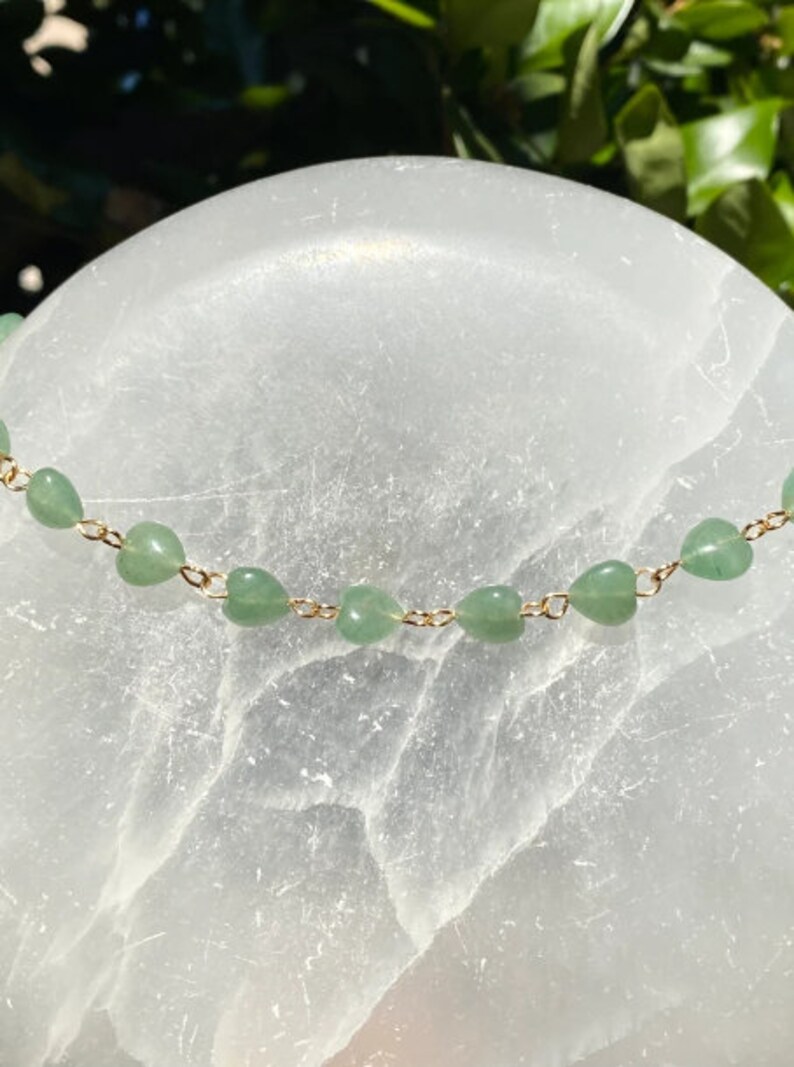 Pu&ograve; includere: Una delicata collana con piccole perline verdi a forma di cuore infilate su una catena color oro. La collana &egrave; esposta su uno sfondo bianco strutturato, con una luce soffusa.