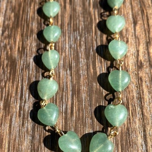 Pu&ograve; includere: Una delicata collana con perline verdi a forma di cuore unite da maglie di catena color oro. Le perline hanno un aspetto liscio e lucido. La collana &egrave; esposta su una superficie di legno.