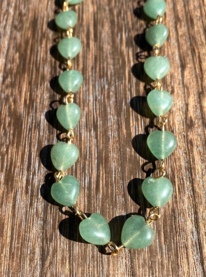 Pu&ograve; includere: Una delicata collana con perline verdi a forma di cuore unite da maglie di catena color oro. Le perline hanno un aspetto liscio e lucido. La collana &egrave; esposta su una superficie di legno.