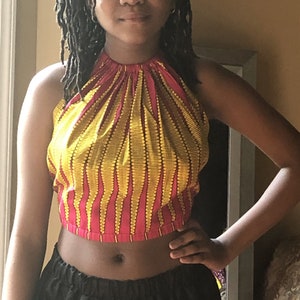 May include: A halter top with a vibrant pattern of yellow vertical lines on a pink background. The top is a crop top style, and the fabric appears to be cotton. The top is paired with black bottoms.