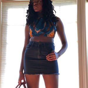 May include: A sleeveless crop top featuring a vibrant blue and orange geometric pattern paired with a dark blue denim mini skirt with a frayed hemline. The outfit is accessorized with round gold-rimmed glasses and a structured woven handbag.