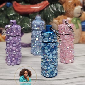 May include: Four small, decorative containers with lids. Each container is covered in iridescent beads and rhinestones. The containers are in shades of purple, blue, and pink. The containers are arranged in a row on a wooden surface.