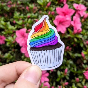 May include: A sticker of a cupcake with rainbow frosting. The cupcake has a brown base and a white liner with vertical lines. The frosting is a swirl of red, orange, yellow, green, blue, and purple.