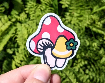 May include: A colorful sticker featuring a whimsical mushroom design. The mushroom has a red cap with white polka dots, a yellow stem, and a green flower accent. The sticker is held against a backdrop of green foliage.