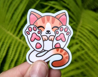 May include: A sticker featuring a cartoon cat with orange and white fur, pink ears, and pink paw pads. The cat is smiling and has its paws up. The sticker has a white border and is held by a hand.