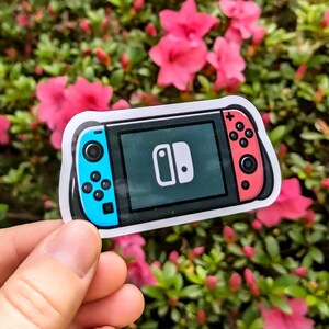May include: A sticker of a video game console with a black screen and blue and red controllers. The console has a white border and a white logo on the screen. The sticker is held in front of a background of pink flowers and green leaves.