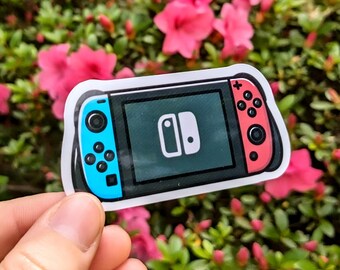 May include: A sticker of a video game console with a black screen and blue and red controllers. The console has a white border and a white logo on the screen. The sticker is held in front of a background of pink flowers and green leaves.