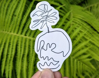 May include: A white sticker featuring a black line drawing of a plant in a pot. The plant has a large, stylized leaf and a wavy base. The sticker is held against a backdrop of green fern leaves.