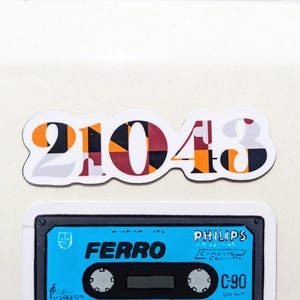 May include: A sticker with the numbers "21043" in a bold font, featuring a pattern of orange, black, and burgundy geometric shapes. Below, a blue cassette tape with the word "FERRO" and "PHILIPS" printed on it.