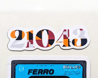 May include: A sticker with the numbers "21043" in a bold font, featuring a pattern of orange, black, and burgundy geometric shapes. Below, a blue cassette tape with the word "FERRO" and "PHILIPS" printed on it.