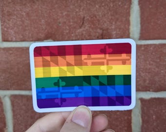 May include: A rectangular sticker with a rainbow flag design. The flag features a gradient of colors, including red, orange, yellow, green, blue, and purple. The sticker also has a cross design in the center.