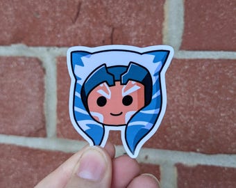 May include: A sticker featuring a cartoon illustration of a character with blue and white markings and a smiling face. The character has blue head tails and is set against a white background. The sticker is held in front of a brick wall.