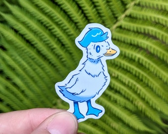 May include: A sticker featuring a cartoon duck wearing a blue hat. The duck is primarily light blue with darker blue accents and a yellow beak. The sticker is held against a backdrop of green fern leaves.
