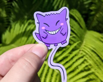 May include: A purple sticker featuring a cartoon character with a wide grin and pink cheeks. The character has a jagged outline and a long, curved tail. The sticker is held against a backdrop of green foliage.