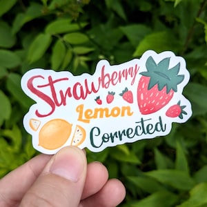 May include: A white sticker with the words "Strawberry Lemon Corrected" in red, yellow, and green. The sticker features illustrations of strawberries and a lemon. The sticker is held in front of a green leafy background.