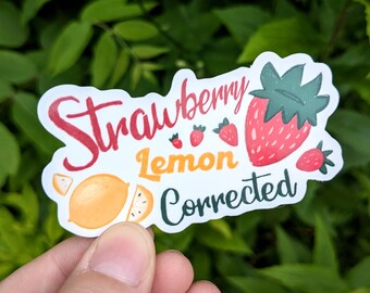 May include: A white sticker with the words "Strawberry Lemon Corrected" in red, yellow, and green. The sticker features illustrations of strawberries and a lemon. The sticker is held in front of a green leafy background.