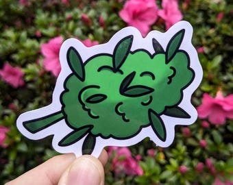 May include: A green sticker featuring a cartoon illustration of a cannabis bud with dark green leaves and a white border. The sticker is held in a hand against a background of pink flowers and green foliage.