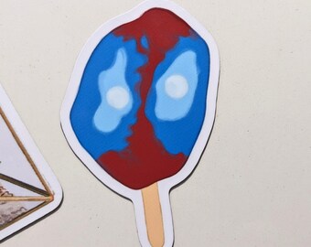 May include: A sticker in the shape of a popsicle with a blue and red design. The popsicle has a white outline and a light brown stick. The blue section has two white circles.