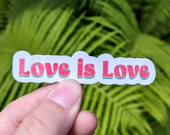 May include: A sticker with the phrase "Love is Love" in red, outlined in blue and yellow, on a white background. The sticker is held against a backdrop of green foliage.