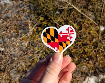 May include: A heart-shaped sticker featuring the Maryland state flag design. The sticker is divided into two halves, one with a black and gold diamond pattern and the other with a red and white cross design. The sticker is held in a hand.