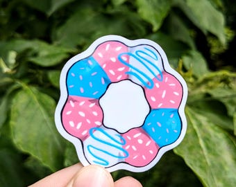 May include: A sticker featuring a stylized donut design. The donut is decorated with pink and blue sections, white sprinkles, and blue icing swirls. The sticker has a white border and a white center.