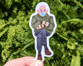May include: A sticker featuring an illustration of a person wearing a face mask, green jacket, and patterned mittens. The person is seated with legs crossed, wearing dark pants and brown shoes. The background is a blurred green.