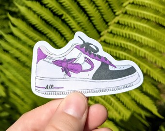 May include: A sticker of a white sneaker with purple accents, black laces, and a black toe. The word "AIR" is printed on the side of the sole. The sneaker has a purple swoosh and a purple flower design.
