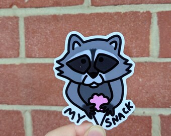 May include: A sticker featuring a cartoon raccoon holding a pink ice cream cone. The raccoon is gray and white with black eyes and a black nose. The text "MY SNACK" is written in black on the sticker.