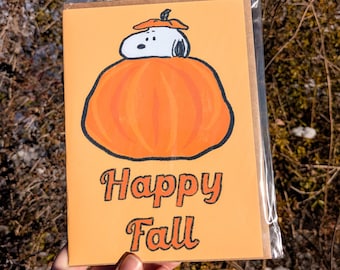 May include: Greeting card featuring Snoopy from Peanuts, atop a large orange pumpkin. The card has the words "Happy Fall" in orange and black lettering on a light orange background. The card is in a clear plastic sleeve.