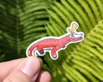 May include: A red and white sticker featuring a cartoon-style lizard with yellow accents. The lizard has a long body, a white skull-shaped head, and is set against a blurred green background of fern leaves.