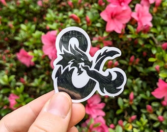 May include: A black and white sticker featuring a dragon design integrated into an ampersand symbol. The sticker is held against a backdrop of green foliage and pink flowers, creating a vibrant contrast.
