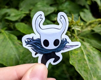 May include: A white and black sticker featuring a stylized character with a horned head, two large black eyes, and a flowing blue cape. The character is holding a silver blade. The sticker is held in a hand against a green leafy background.