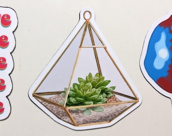 May include: A sticker featuring a geometric terrarium with a succulent plant inside. The terrarium has a gold-colored frame and clear glass panels. The succulent is green and the soil is brown. The sticker is white with a black outline.