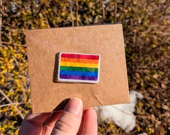 May include: A rectangular pin featuring the rainbow colors of the Pride flag. The pin is set against a brown cardboard backing. The pin has a white border and is held by a hand.