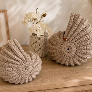 Könnte beinhalten: Zwei beige gehäkelte Körbe mit einem muschelartigen Spiraldesign. Jeder Korb besteht aus dickem, strukturiertem Garn mit einer kreisförmigen Mitte und einer erhöhten, geschwungenen Haube. Ein Korb hat eine kleine braune Lederlasche mit Niete.