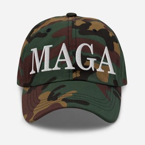Op de afbeelding: Een camouflage baseballpet met de witte geborduurde letters "MAGA" over de voorkant. De pet heeft een klassiek zes-panelen ontwerp met een gebogen klep en een verstelbare band aan de achterkant.