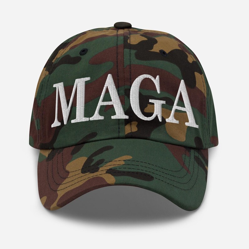Op de afbeelding: Een camouflage baseballpet met de witte geborduurde letters "MAGA" over de voorkant. De pet heeft een klassiek zes-panelen ontwerp met een gebogen klep en een verstelbare band aan de achterkant.