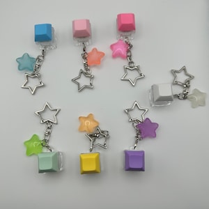 May include: A collection of colorful keycap earrings. Each earring features a square keycap in various colors like blue, pink, and yellow, paired with star-shaped charms in coordinating hues. The earrings are connected by silver-toned chains.