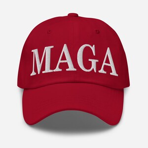 Op de afbeelding: Rode baseballcap met de witte geborduurde letters "MAGA" op de voorkant. De cap heeft een gebogen klep en een stoffen bandsluiting aan de achterkant.
