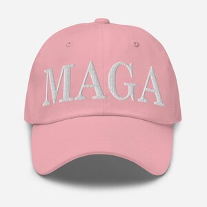 Op de afbeelding: Een lichtroze baseballpet met de wit geborduurde letters "MAGA" op de voorkant. De pet heeft een gebogen klep en een verstelbare stoffen band aan de achterkant.