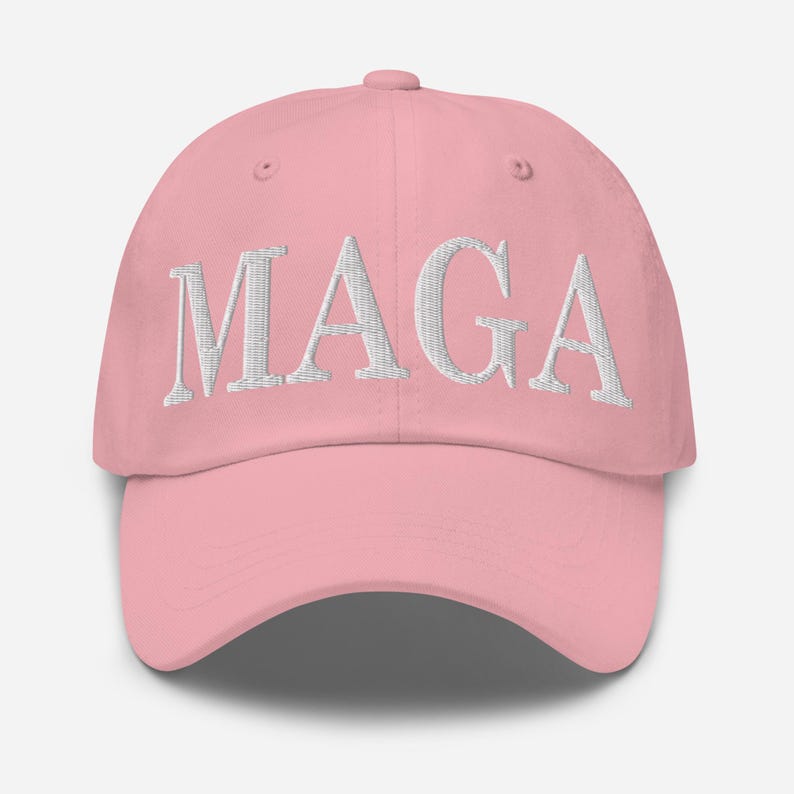 Op de afbeelding: Een lichtroze baseballpet met de wit geborduurde letters "MAGA" op de voorkant. De pet heeft een gebogen klep en een verstelbare stoffen band aan de achterkant.