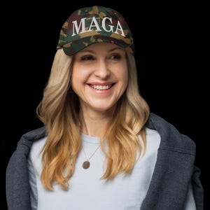 Op de afbeelding: Een camouflage baseballpet met het woord "MAGA" in het wit geborduurd. De pet wordt gedragen door een persoon met blond haar, die een lichtblauw shirt en een grijze jas draagt.