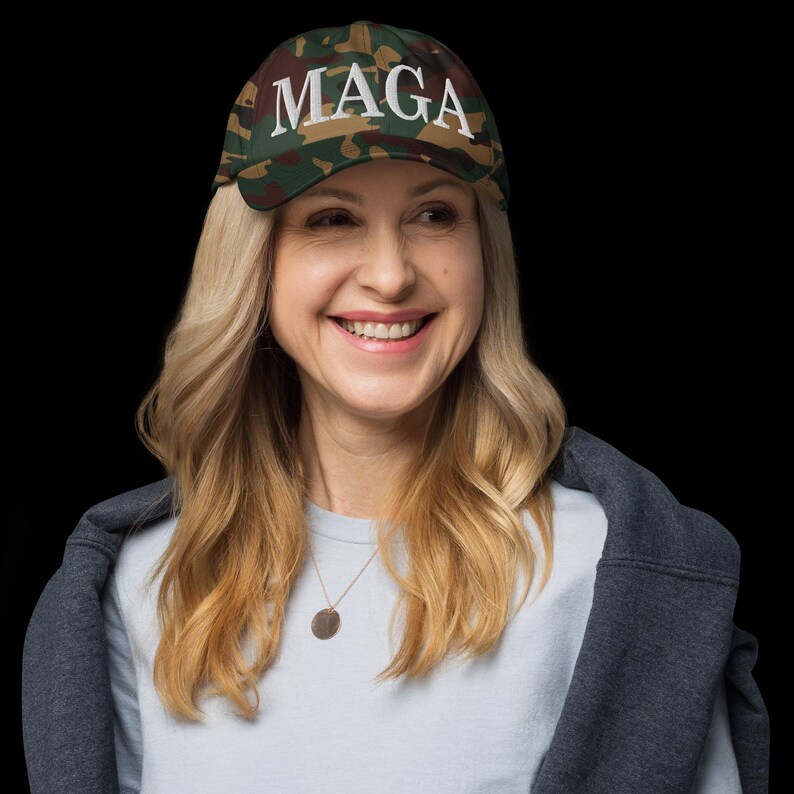Op de afbeelding: Een camouflage baseballpet met het woord "MAGA" in het wit geborduurd. De pet wordt gedragen door een persoon met blond haar, die een lichtblauw shirt en een grijze jas draagt.