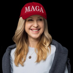 Op de afbeelding: Een rode baseballpet met de wit geborduurde letters "MAGA". De pet wordt gedragen door een persoon met blond haar, die een lichtblauw shirt en een grijze jas draagt. De achtergrond is zwart.