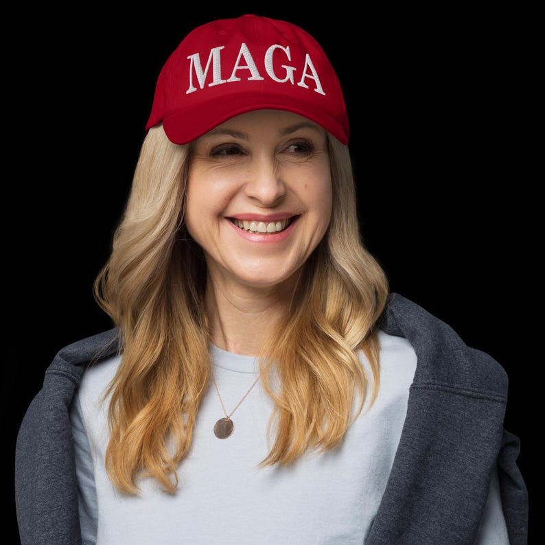 Op de afbeelding: Een rode baseballpet met de wit geborduurde letters "MAGA". De pet wordt gedragen door een persoon met blond haar, die een lichtblauw shirt en een grijze jas draagt. De achtergrond is zwart.
