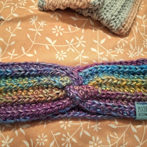 Puede incluir: Una diadema de crochet multicolor con un diseño anudado. La diadema tiene un esquema de color azul, morado, amarillo y verde. Una pequeña etiqueta azul con el texto "MADE WITH ♥" está unida a la diadema.